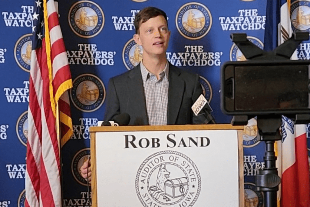 Rob Sand behind a podium which has his name on it and the seal for the auditor's office. He stands in front of a blue backdrop with an alternating pattern of the auditor seal and the words "The Taxpayers' Watchdog"